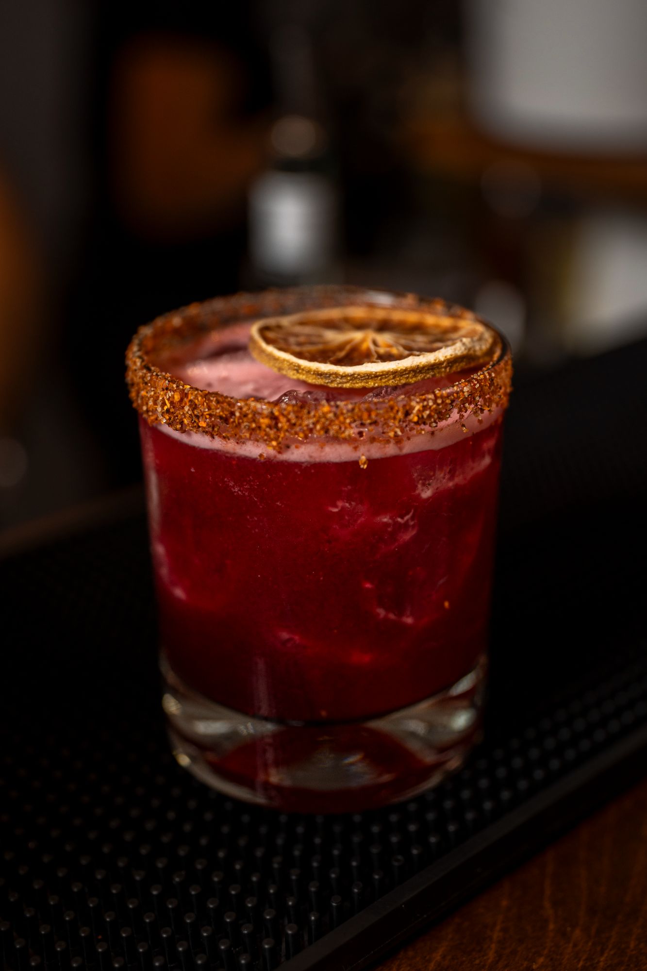 A deep red hibiscus cooler with a chile-salt rim and a dried citrus wheel on a dark bar mat