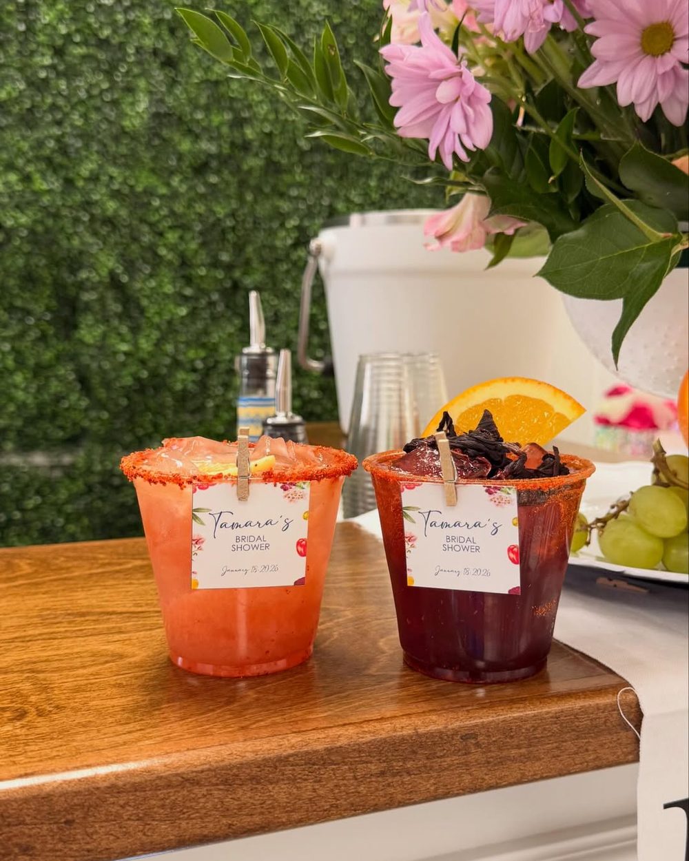 Two chamoy-rimmed cocktails with printed tags clipped to the cups, against a boxwood wall