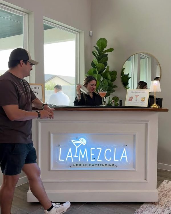 A bartender shaking a cocktail behind the lit La Mezcla bar while a guest waits