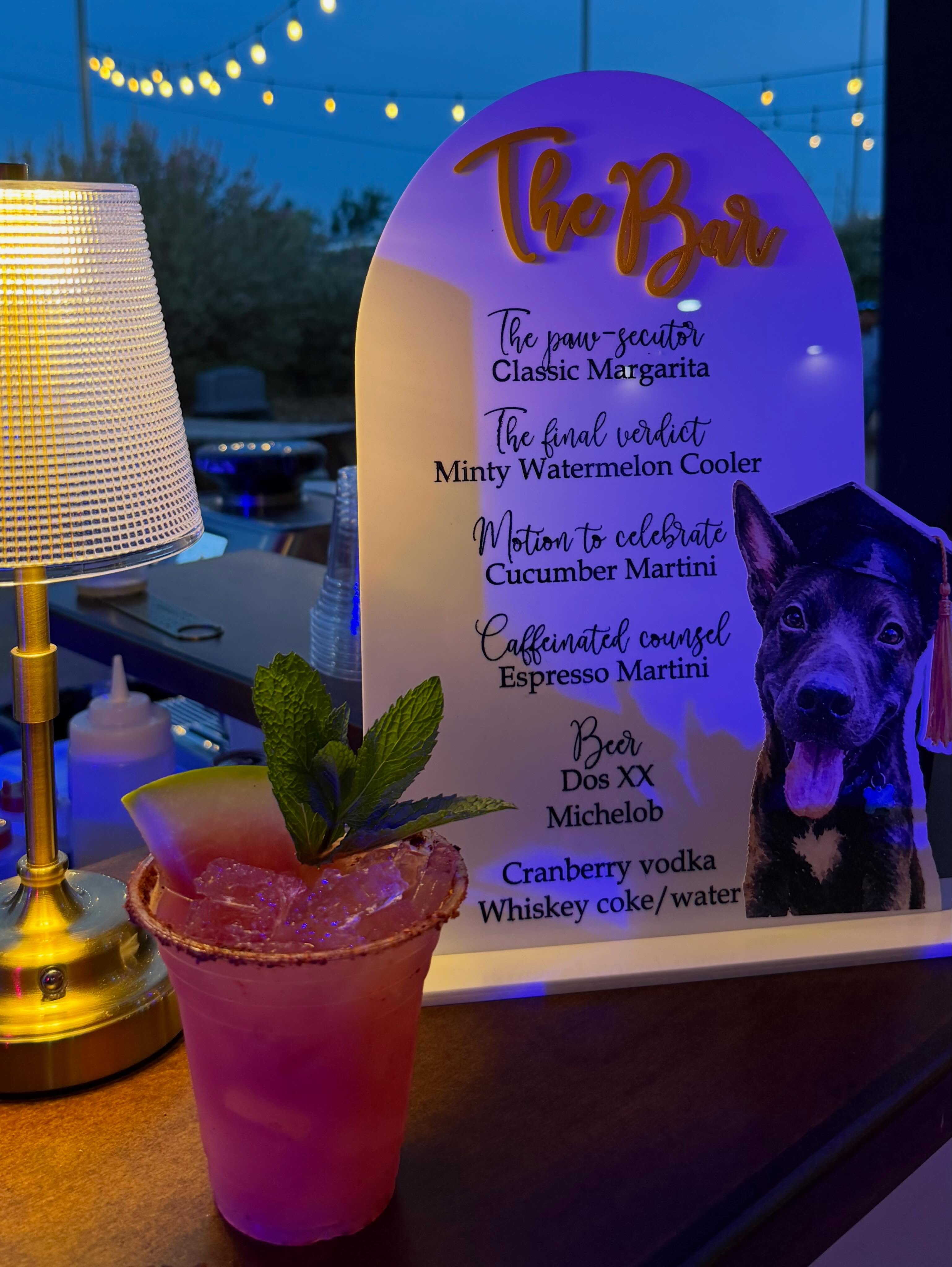 An arched bar menu at dusk with every drink renamed for the law graduate — the paw-secutor, the final verdict, motion to celebrate — beside a cut-out of the family dog in a graduation cap, with a chile-rimmed watermelon cooler garnished with mint in front and string lights behind