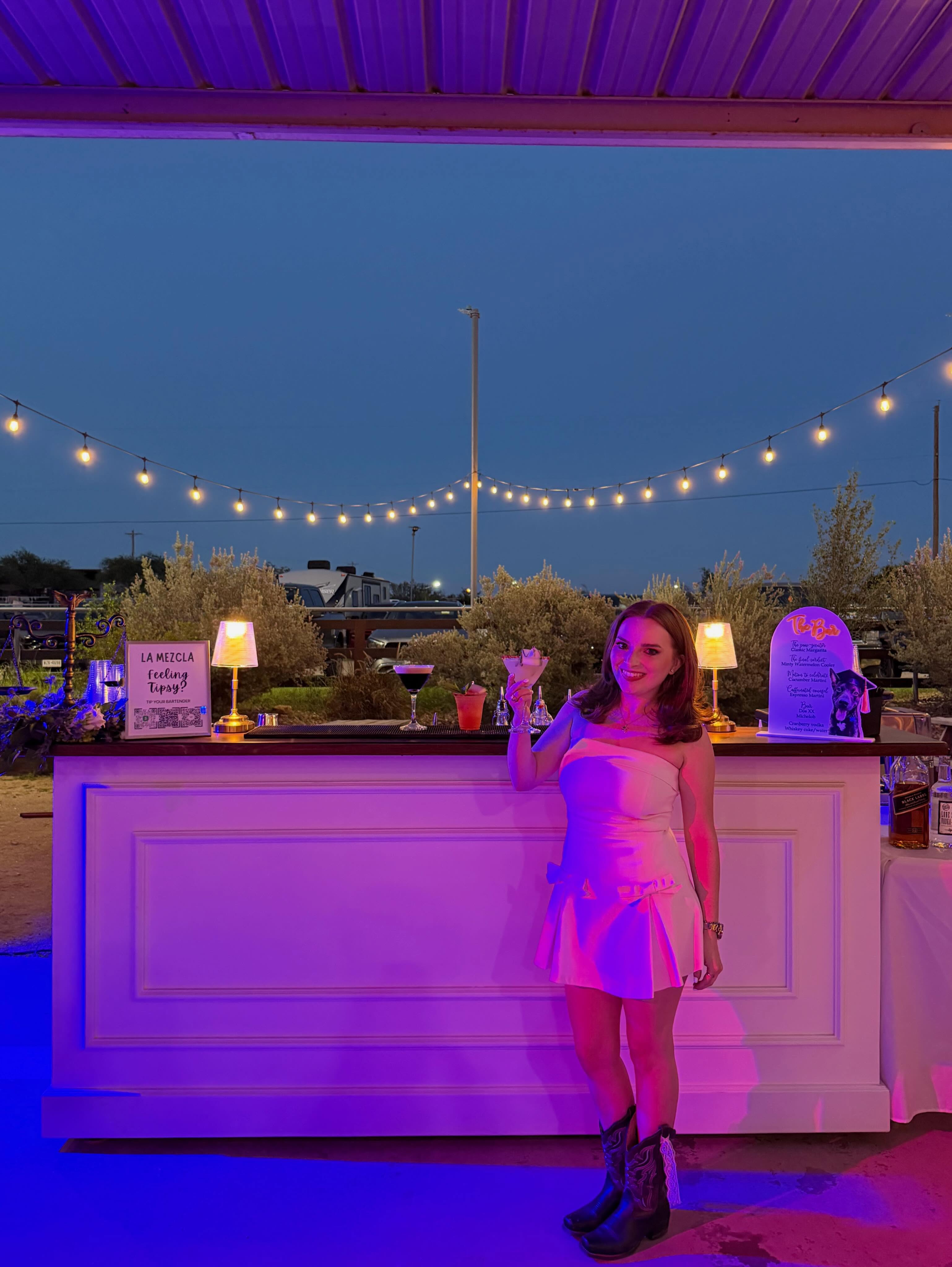 A guest in a white dress and cowboy boots raising a coupe beside the La Mezcla bar, lit pink and purple under string lights against a deep blue dusk sky