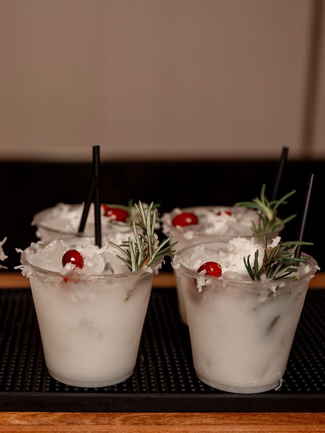 A framed Signature Drinks card describing the White Xmas Margarita and the Spiced Cranberry Paloma, standing on the bar beside both cocktails