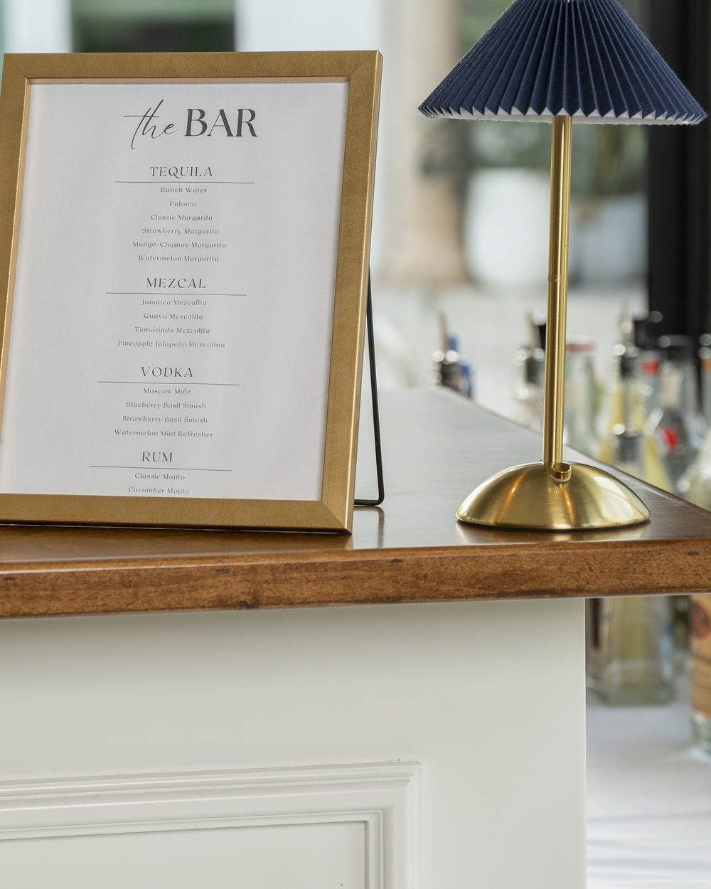 A framed bar menu listing tequila, mezcal, vodka and rum, beside a pleated lamp