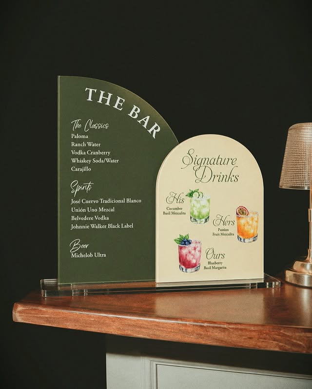 Two arched menu boards on the bar — a green The Bar card listing the classics, spirits and beer, beside a cream Signature Drinks card naming His, Hers and Ours with a painting of each drink