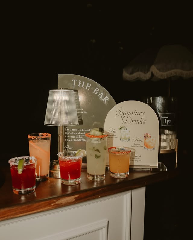 Six cocktails in a row along the bar top in front of the arched bar and signature drinks menus, lit by a small crystal lamp