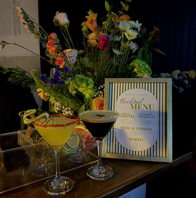 A chile-rimmed cucumber martini and an espresso martini topped with coffee beans, standing on the bar in front of the framed Tini Bit Older cocktail menu with a tall arrangement of roses, lilies and hydrangea behind