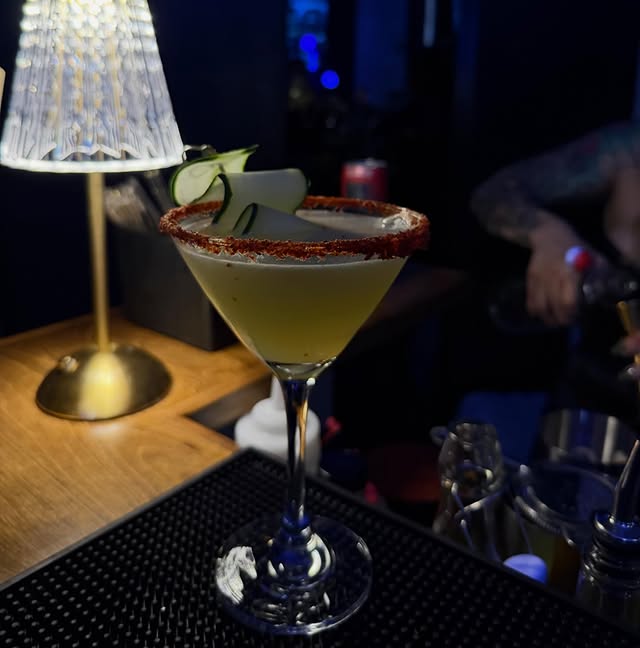 A cucumber martini with a chile-salt rim and a folded ribbon of cucumber, on a black bar mat with the brass lamp glowing behind and a bartender working out of focus