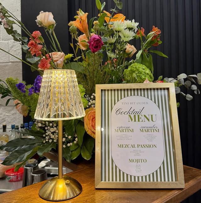 A gold-framed cocktail menu headed Tini Bit Older listing espresso martini, cucumber martini, mezcal passion and mojito, lit by a small brass lamp with a fluted crystal shade against a bank of flowers