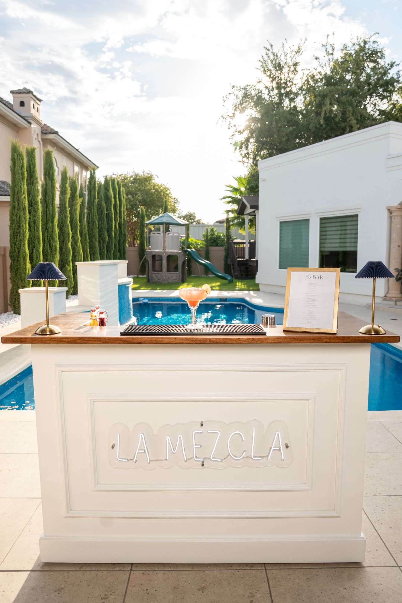 The white La Mezcla bar with its neon sign lit, set up beside a blue swimming pool with a framed bar menu, two pleated lamps and a margarita on the wooden top