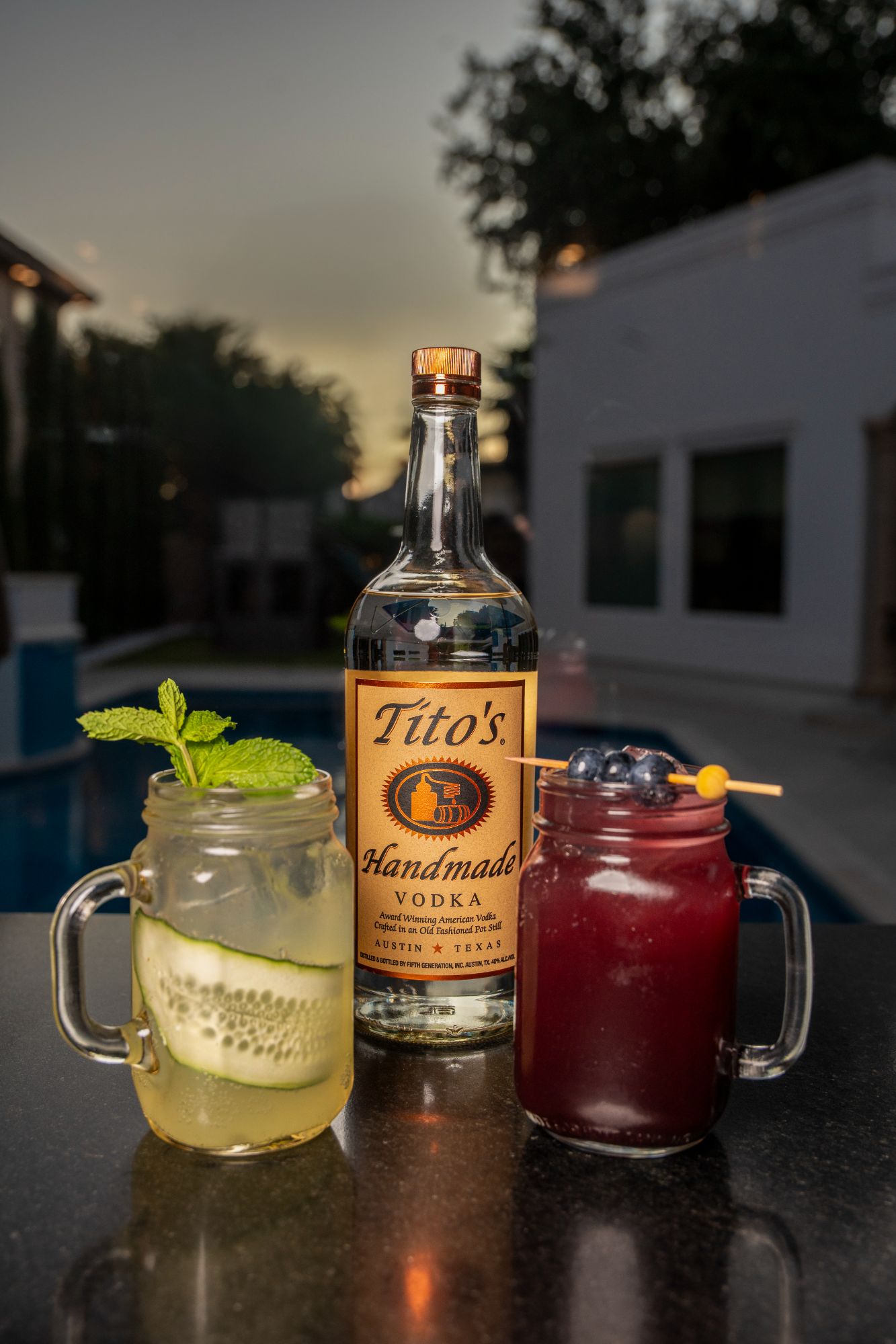 Cucumber mint and blueberry basil cocktails in mason jar mugs beside a vodka bottle at dusk